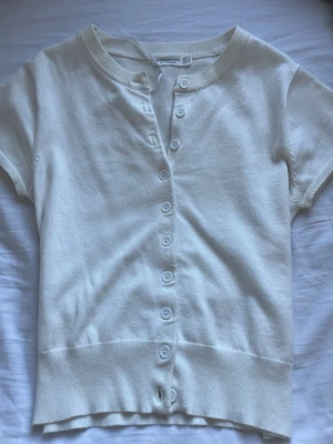 Vit kortärmad topp med knappar Uniqlo - Söt vit kofta från Stradivarius med korta ärmar  Perfekt till både vardag och sommar! Koftan är i fint skick. Den har en liten fläck längst ut i kanten (se sista bilden), men den är knappt märkbar och påverkar inte helhetsintrycket. Superfin och lätt att matcha – säljs då den tyvärr inte kommer till användning längre 