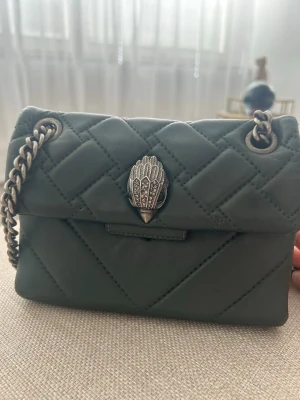 Grön quiltad handväska med kedja - Snygg mörkgrön quiltad handväska i mjukt läder med silverfärgad kedjerem och dekorativt spänne framtill. Väskan har en kompakt rektangulär form och cool detalj med metallspänne i form av en hand. Perfekt accessoar för att lyfta din outfit. Ett litet hål inne i väskan men som inte syns alls!