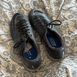 Nike Mercurial fotbollsskor svarta/blå inhomhus - Säljer ett par Nike Mercurial fotbollsskor i svart med blå och silverdetaljer. Skorna har snörning, låg profil och yttersula anpassad för snabba rörelser på planen. Materialet är syntetiskt och designen är sportig och modern med tydliga Nike-loggor.