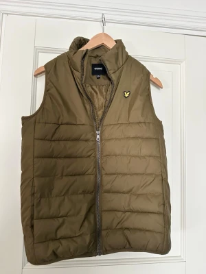 Olivgrön dunväst från Lyle & Scott - Snygg olivgrön dunväst från Lyle & Scott med gul logga på bröstet. Västen har hög krage, dragkedja framtill och quiltad design. Perfekt att slänga över en hoodie eller tröja för extra värme. Materialet är lätt och smidigt.