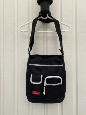 Svart axelväska från nypd  - Axelväska från nypd med justerbar axelrem. Väskan har flera praktiska fack framtill, vita detaljer och dragkedja. I frächt skick trots åldern. Mäter och fler bilder vid intresse. Vintage stuk! Djur och rökfritt hem 👍