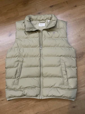 Beige dunväst från Gant - Snygg beige dunväst från Gant med hög krage och dragkedja framtill. Västen har två fickor med dragkedja och Gant-logga med sköld på bröstet. Perfekt för lager-på-lager och riktigt skön att bära under kyliga dagar.