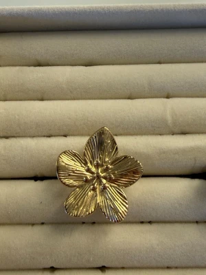 Guldfärgad blomformad ring - Unik ring i guldfärg med en stor blomformad design. Ringen har fem detaljerade kronblad med räfflade ytor och små guldfärgade kulor i mitten. Perfekt för dig som vill sticka ut med ett statement-smycke.
