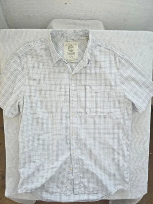 Vit rutig skjorta från Timberland - Klassisk vit kortärmad skjorta från Timberland med diskret rutigt mönster i blått och grått. Skjortan har en bröstficka, knappar framtill och en avslappnad regular fit. Perfekt till jeans eller shorts för en clean och enkel stil.