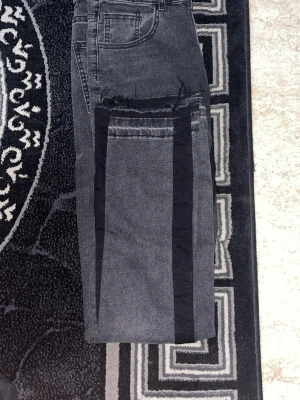 Grå jeans med svarta detaljer - Säljer ett par grå jeans med svarta vertikala ränder längs benen och råa kanter vid fickorna. Jeansen har klassisk femficksmodell och raka ben. Perfekta för dig som vill ha en unik och trendig look.