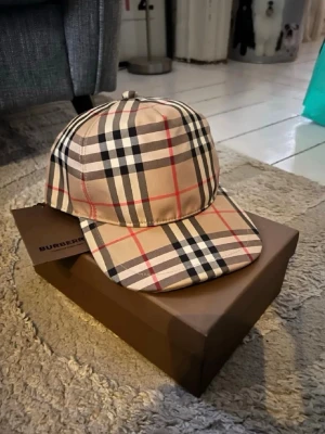 Burberry klassisk rutig keps - Säljer en ikonisk Burberry keps i beige med det klassiska rutmönstret i svart, vitt och rött. Kepsen har böjd skärm och justerbar snapback baktill. Märkt med Burberry London England på baksidan. Perfekt accessoar för en trendig och stilren look.