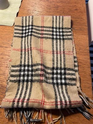 Beige rutig halsduk från Burberrys - Klassisk beige halsduk från Burberrys i 100% lammull med det ikoniska rutmönstret i svart, vitt och rött. Halsduken har fransar i ändarna och är tillverkad i England. Perfekt accessoar för att lyfta din outfit med en tidlös look.