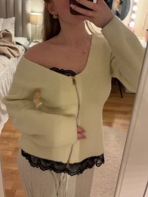 Benvit stickad kofta från mango - Trendig ljusgul stickad kofta med dragkedja framtill och vida ärmar. Koftan har en snygg off-shoulder passform och svart spetsdetalj längst ner. Perfekt för dig som vill ha en unik och stilren look. Nypris 600kr