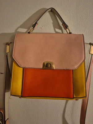 Flerfärgad axelväska med guldfärgade detaljer - Handväska i färgerna rosa, mörk orange och gul. Väskan har ett lock med guldfärgat spänne, justerbar axelrem och ett topphandtag. Tillverkad i slätt konstläder med stilren, fyrkantig form och färgblockad design. Metallerna har slitningar och insidan är lite smutsig, annars bra skick.