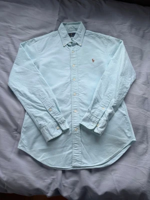 Ljusblå skjorta från Ralph Lauren - Klassisk ljusblå skjorta från Ralph Lauren med broderad logga på bröstet. Skjortan har knappar framtill, lång ärm och button-down krage. Tillverkad i mjuk bomull för en clean och stilren look.