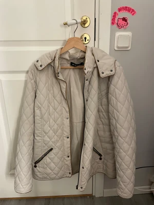 Beige quiltad jacka från Micha - Snygg beige quiltad jacka från Micha med dragkedja och knappar framtill. Jackan har två fickor med dragkedja och en avtagbar huva. Perfekt för kyligare dagar och har en klassisk, tidlös look.