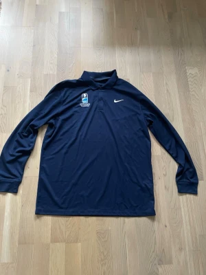 Mörkblå långärmad skjorta Nike - Mörkblå långärmad skjorta från Nike med klassisk krage och knappar framtill. Skjortan har ett broderat Nike-märke på bröstet och en sportig logga på vänster sida. Tillverkad i ett mjukt och funktionellt material som passar perfekt till sportiga tillfällen.