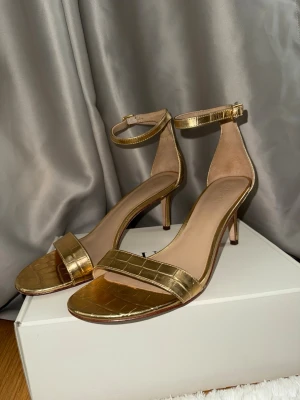Guldiga sandaletter från Ralph Lauren - Säljer ett par sandaletter i guld från Ralph Lauren. De har justerbar ankelrem och låg klack. Ytan har ett croco-mönster och materialet är läder. Storleken är EU 40 cirka 26 cm när jag mätte insidan fram till bak Nypris: 1895 kr
