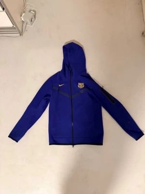 Blå Nike FC Barcelona hoodie - Snygg blå hoodie från Nike med FC Barcelona-logga på bröstet och dragkedja framtill. Tröjan har huva, svarta detaljer och en ficka på ärmen. Perfekt för dig som älskar fotboll och vill ha en sportig look.