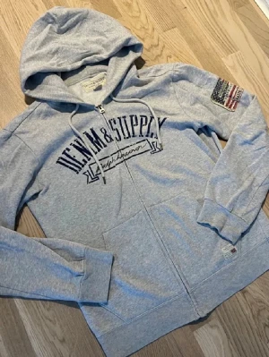 Grå hoodie från Ralph Lauren - Snygg grå zip-hoodie från Ralph Lauren Denim & Supply med tryck på bröstet och en patch med amerikanska flaggan på ärmen.
