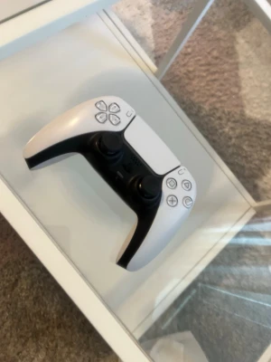 Vit Sony DualSense PS5 handkontroll - Snygg vit DualSense handkontroll till PlayStation 5 från Sony. Kontrollern har svart mittparti, två analoga spakar, touchpad och klassiska PlayStation-knappar. Ergonomisk form och tillverkad i plast, perfekt för gaming och långa spelsessioner.