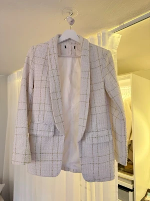 Vit rutig kavaj i bouclétyg - Stilren vit kavaj i bouclétyg med rutigt mönster i beige och guld. Kavajen har två fickor framtill med lock och öppen framsida utan knappar. Perfekt för att lyfta en outfit med en elegant touch.