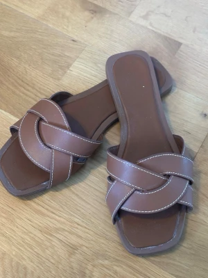 Bruna sandaler med flätad rem - Snygga bruna sandaler med breda, flätade remmar i konstläder och vita sömmar. De har fyrkantig tå och låg sula, perfekt för en avslappnad och trendig look. Passar till jeans, kjol eller shorts för en chill vibe.