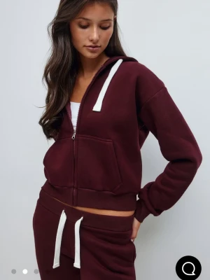 Vinröd hoodie med dragkedja - Vinröd hoodie med dragkedja och vita snören i huvan. Modellen har en croppad passform, känguruficka framtill och ribbade muddar. Perfekt för en avslappnad och trendig look. Tillverkad i mjukt sweatshirtmaterial.