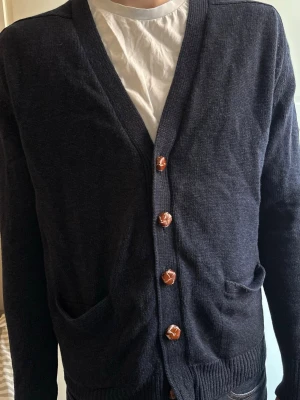 Mörkblå kofta från Barbour - Snygg mörkblå kofta med v-ringning och fyra bruna knappar framtill. Koftan har två fickor och ribbade muddar vid ärmslut och nederkant. Perfekt att slänga över en t-shirt för en chill look.