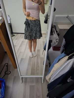 Svart blommig kortkjol med knyt - Säljer en svart kortkjol med vitt blommönster och knyt i midjan. Kjolen har en avslappnad passform och är perfekt för varmare dagar. Materialet känns lätt och flowy, vilket gör den superbekväm att bära. Använd endast nån gång.