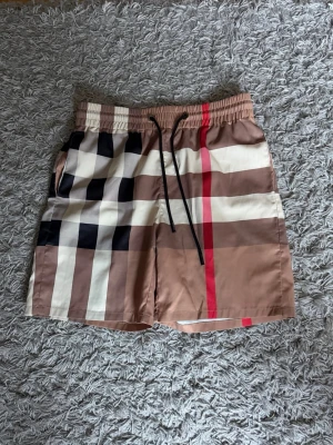 Burberry badbyxor med rutigt mönster - Säljer ett par snygga Burberry badbyxor i klassiskt beige, brunt, svart och vitt rutmönster med en röd rand. De har resår i midjan, svarta dragsnören och meshfoder på insidan. Perfekta för stranden eller poolen!