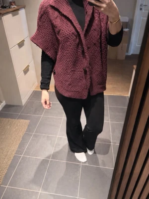 Vinröd stickad poncho från Soaked - Vinröd stickad poncho från Soaked med grovstickad struktur och dekorativa flätor. Ponchon har en hög krage och stora svarta knappar längs sidorna. Modellen är loose fit och har en rundad form som ger en chill vibe. Perfekt att slänga över en långärmad tröja för extra värme. Saknar dock en knapp.