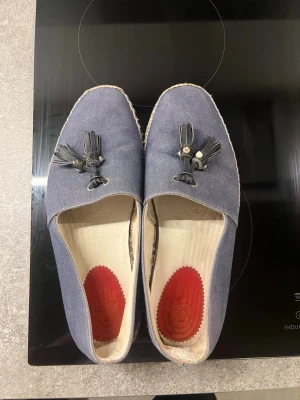 Blå loafers från Christian Louboutin - Blå loafers från Christian Louboutin i canvas med dekorativa tofsar och detaljer på ovandelen. Skorna har den ikoniska röda sulan och insida i ljust läder med röd logga vid hälen. Perfekta för dig som vill sticka ut med stil.