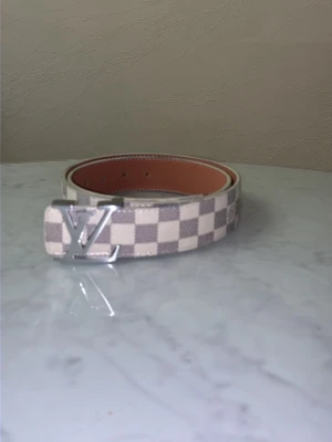 Rutigt bälte - Snyggt bälte i Damier Azur-mönster med vita och gråa rutor. Bältet har en silvrig logga som spänne och insidan är ljusbrun. Perfekt accessoar för att lyfta din outfit med en lyxig touch.