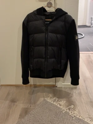 Svart Bvaldi cardigan - Snygg svart Bvaldi cardigan med huva och dragkedja framtill. Jackan har vadderad kropp i glansigt material och stickade ärmar med bavldi logga på vänster ärm. Perfekt för dig som vill ha en riktigt trendig look.