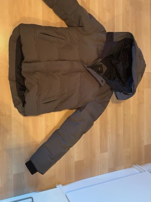 Grå dunjacka från Canada Goose - Säljer en brun dunjacka från Canada Goose i storlek M. Jackan har huva, två snedställda fickor framtill och breda muddar vid ärmsluten. Perfekt för kalla vinterdagar och riktigt varm tack vare dunfyllningen.