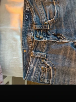 Blå bootcut jeans från Miss me - Snygga blå bootcut jeans från Miss me  med slitningar och detaljerade sömmar. Jeansen har dubbla knappar i midjan, dekorativa nitar och klassiska fem fickor. Materialet är denim med en tvättad look och lätt slitna partier för en trendig vibe. De saknas en knapp på bild 3, men inget man märker. Köpte på Vinted. Bra kvalitet. Passar xs-s!! pris kan diskuteras men inte för mycket!!