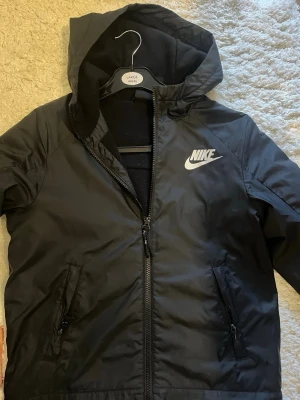 Svart Nike vindjacka med huva - Snygg svart vindjacka från Nike med dragkedja framtill och två fickor med dragkedja. Jackan har en praktisk huva och Nike-logga i vitt på bröstet. Tillverkad i ett lätt och tåligt material som skyddar mot vind.