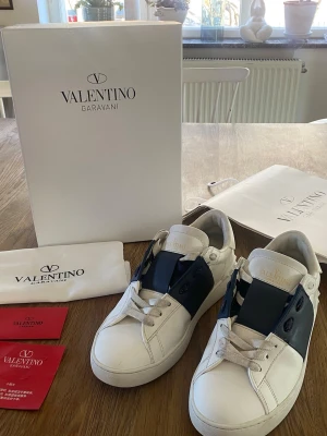 Valentino Garavani vita sneakers - Snygga vita sneakers från Valentino Garavani med bred blå läderdetalj över vristen och vita skosnören. Skorna har en klassisk låg siluett och diskret logga på plösen. Tillverkade i läder och levereras med originalkartong och dustbag.