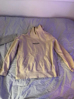 Beige hoodie från Jack & Jones - Säljer en beige hoodie från Jack & Jones Originals med broderad text på bröstet. Hoodien har en stor känguruficka framtill och en klassisk huva med dragsko. Insidan är mjuk och fleecefodrad för extra komfort. Perfekt för chill dagar eller när du vill vara extra bekväm.