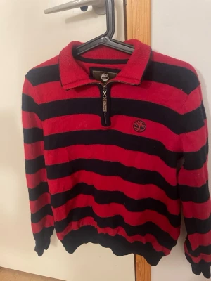 Randig Timberland tröja med dragkedja - Snygg röd och svart randig tröja från Timberland med halv dragkedja och klassisk krage. Tröjan har långa ärmar, ribbade muddar och broderad Timberland-logga på bröstet. Perfekt för dig som gillar streetwear och vill ha en bekväm men stilren look.