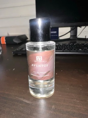 Aventus Eau de Parfum - Säljer en stilren parfymflaska från Inspired By med doften Aventus. Flaskan är genomskinlig med en svart cylinderformad kork och har en brun etikett med vit text. Perfekt för dig som gillar exklusiva och moderna dofter. Cirka 40ml kvar
