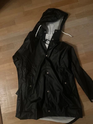 Svart regnjacka med huva - Snygg svart regnjacka med huva och vita dragskor. Jackan har tryckknappar framtill, två fickor med lock och knapp samt justerbara ärmslut. Materialet är glansigt och vattenavvisande, perfekt för regniga dagar.