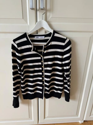 Randig svartvit kofta från Zara - Snygg svartvit randig kofta från Zara med guldfärgade knappar framtill. Koftan har rund halsringning, långa ärmar och ribbade muddar. 