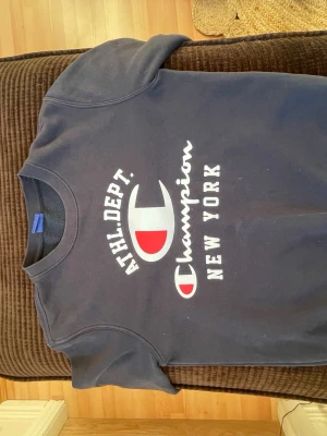 Mörkblå Champion sweatshirt - Snygg mörkblå sweatshirt från Champion med stort vitt och rött tryck på bröstet där det står 'ATHL. DEPT. Champion NEW YORK'. Klassisk rund halsringning och mjukt material, perfekt för en avslappnad streetwear-look.