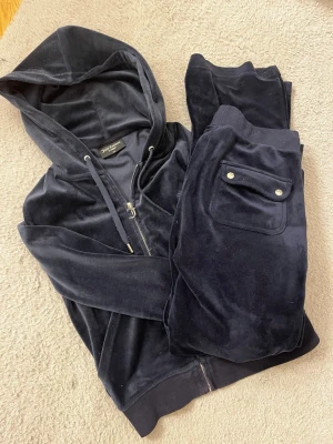 Mörkblå velourset Juicy Couture XS - Säljer ett mörkblått velourset från Juicy Couture i storlek XS. Setet består av en ziphoodie med huva och matchande byxor med ficka bak och silverfärgade detaljer. Perfekt för chill dagar hemma eller när du vill vara extra comfy. Byxor i storlek S