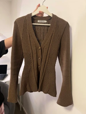 Adoore cardigan  - Snygg brun kofta från Adoore med ribbad struktur och figurnära passform. Koftan har v-ringning, tre knappar framtill och långa ärmar med lätt utsvängd form. Perfekt för lager på lager och enkel att styla till olika looks.