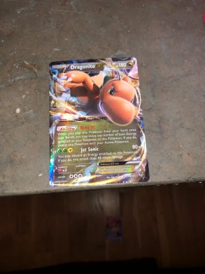 Dragonite EX Pokémonkort - Säljer ett Dragonite EX Pokémonkort med holografisk yta och färgstarkt motiv. Kortet har 180 HP och attacker som Jet Sonic. Texten är på engelska och kortet har en glansig finish med guldfärgade detaljer. Perfekt för samlare eller Pokémonfans.