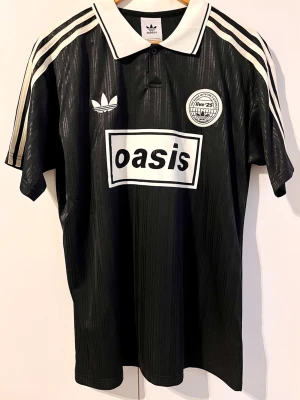 Oasis - Turné 25 - Fotbollströja (M) - Säljer en svart  Oasis-supportertröja för bandets turné under 2025. Tröjan är en reprint i nyskick. Aldrig använd. ”25” på ryggen.      Storlek: Medium