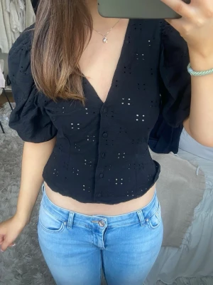 Svart blus med puffärm och v-ringning - Säljer en svart blus med puffärmar och v-ringning. Blusen har broderade detaljer, knappar framtill och är croppad i modellen. Perfekt för dig som gillar en trendig och feminin stil.