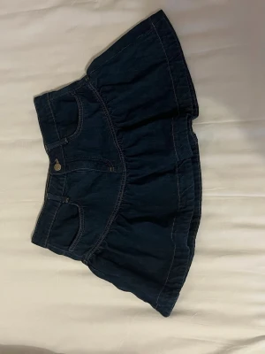 Mörkblå jeanskjol med volang - Säljer en mörkblå kort jeanskjol med volangkant och klassiska fickor fram och bak. Helt oanvänd men har tyvärr ej lapp kvar.