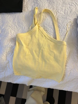 Ljusgul ribbad linnetopp - Snygg ljusgul ribbad linnetopp med smala axelband och asymmetrisk halsringning. Perfekt för varma dagar och enkel att styla med jeans eller kjol. Mjuk och stretchig känsla.