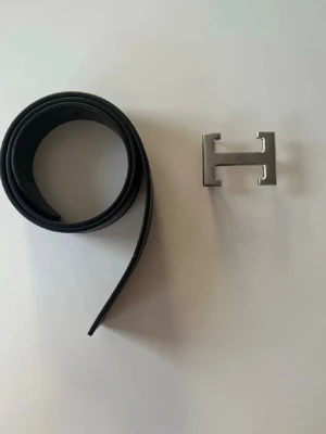 Svart skärp med H-spänne i skinn 115cm - Snyggt svart bälte i skinn med ett stort, silverfärgat H-format spänne. Bältet hakontrastsömmar längs kanterna som ger en clean och modern look. Perfekt accessoar för att lyfta vilken outfit som helst.