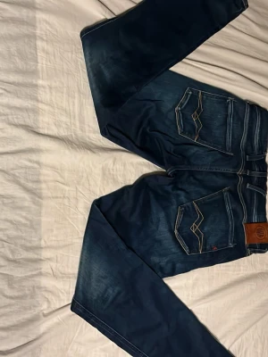 Mörkblå jeans från Replay - Säljer ett par mörkblå jeans från Replay med klassisk femficksdesign och snygga kontrastsömmar. Jeansen har en skinnpatch med Replay-logga bak i midjan och coola detaljer på bakfickorna. Perfekta för dig som gillar en tidlös och trendig look.