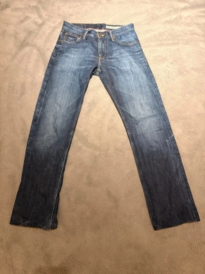 Blå raka jeans från Hilfiger Denim - Snygga blå jeans från Hilfiger Denim med raka ben och klassisk femficksdesign. Jeansen har en lätt tvättad look och kontrastsömmar, samt patch med amerikansk flagga bak i linningen. Perfekta för en avslappnad och trendig stil.